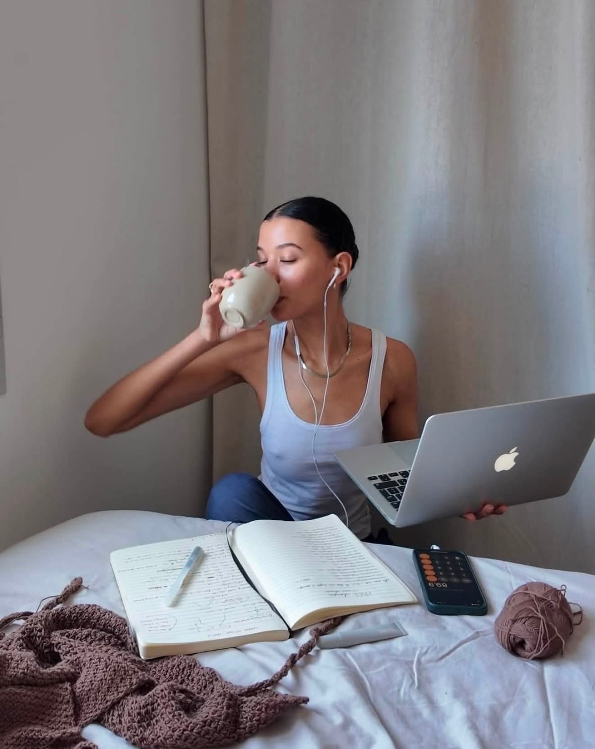 A woman working from bed with a laptop and a mug, notebook and crochet project nearby